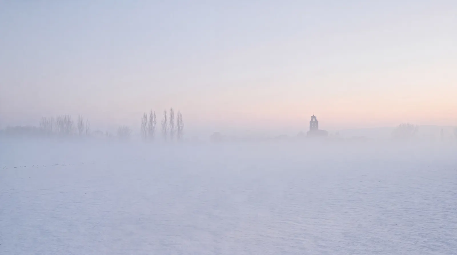 Una llanura nevada vacía al amanecer, con álamos y un pueblo al fondo entre la bruma